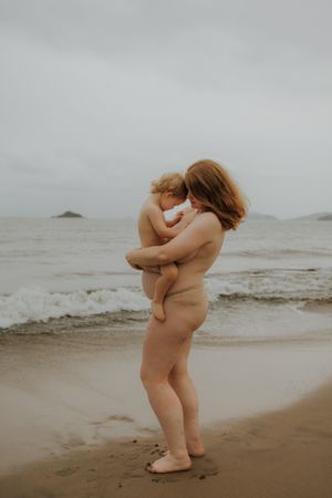 A nude mother tenderly holds her baby on a sandy beach with ocean waves and cloudy skies in the background.