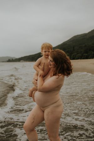Pregnant mother holding toddler on a beach, waves behind them, overcast sky, green hills in background.