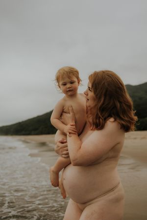 Pregnant woman with red hair holds toddler on beach, waves and green hills in background.