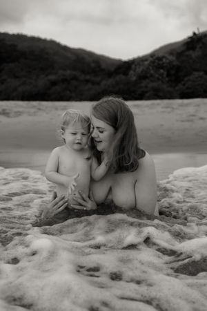 Black and white photo of a mother and toddler playing together on a sandy beach with ocean waves and hills.
