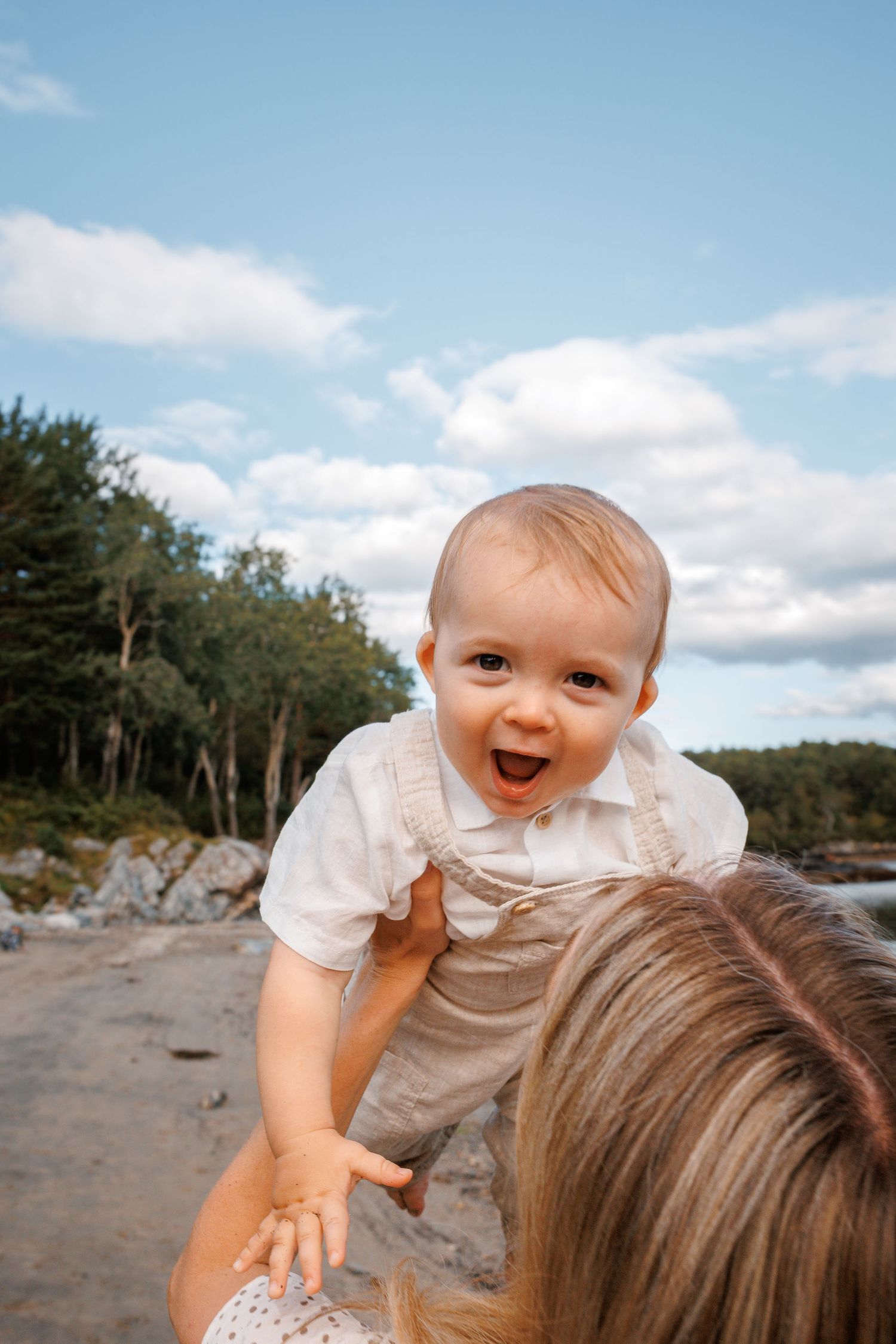 A joyful baby in a white outfit laughs while being held up against a blue summer sky with trees in the background.