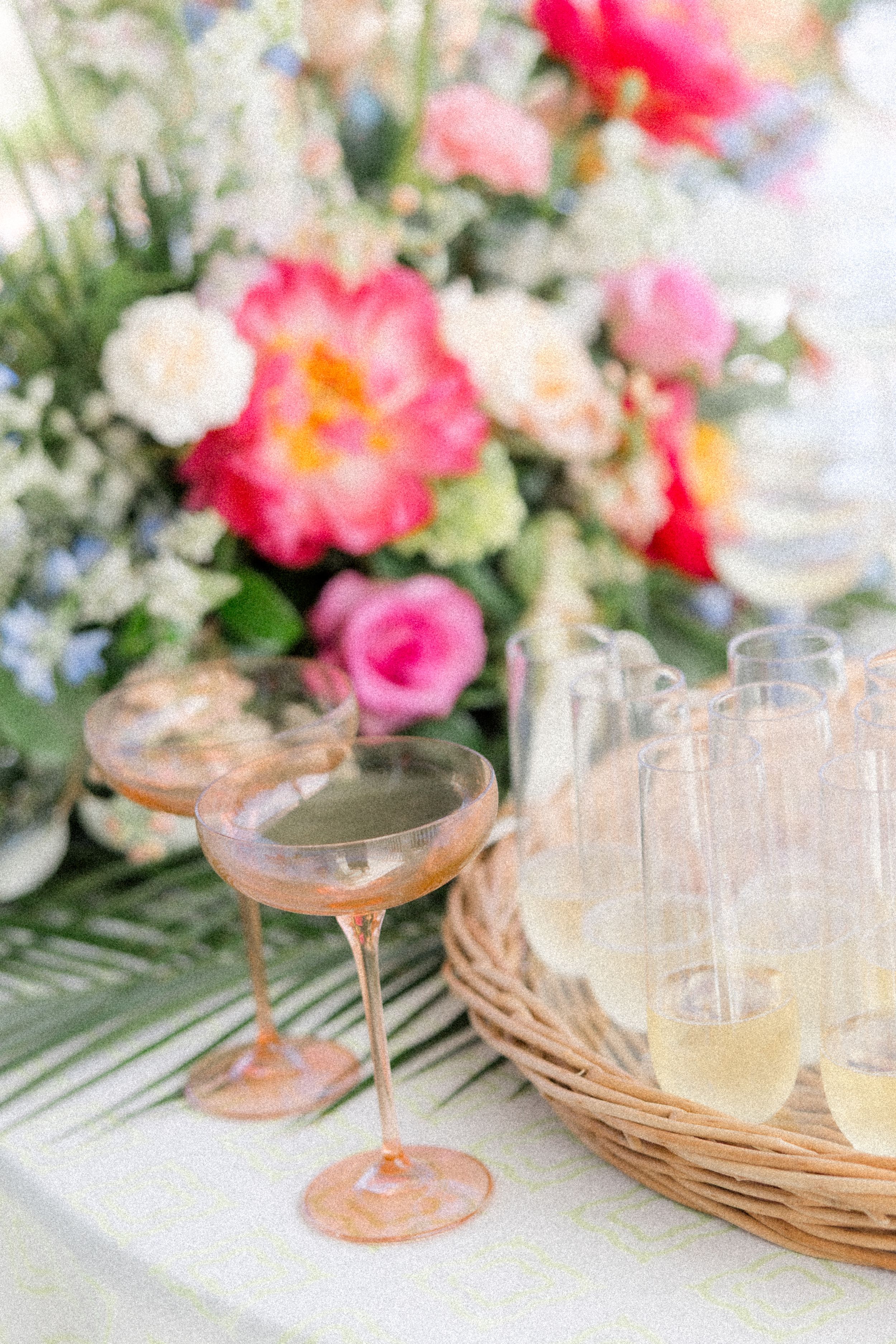 Pink champagne cocktails in coupe glasses arranged on woven chargers with coral flowers and greenery in background.