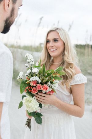A bride in a white dress holds a bouquet of coral roses and white hydrangeas during a beach wedding ceremony.