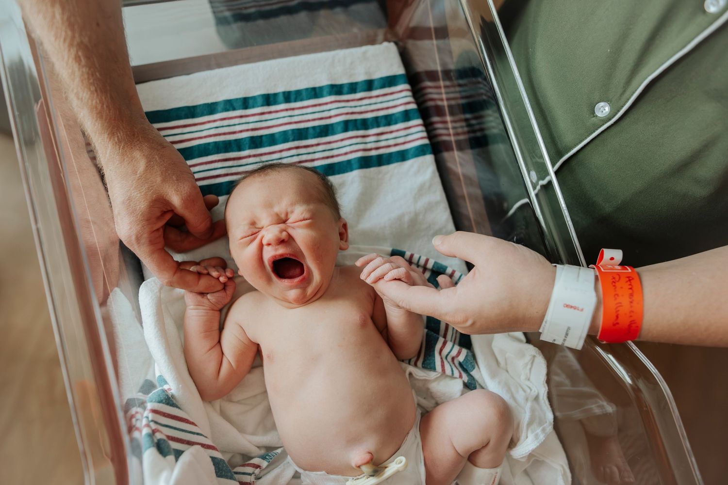 Crying newborn baby lying in hospital bassinet while parents reach in to comfort their new arrival. Ivy Lynn Photography, North Georgia