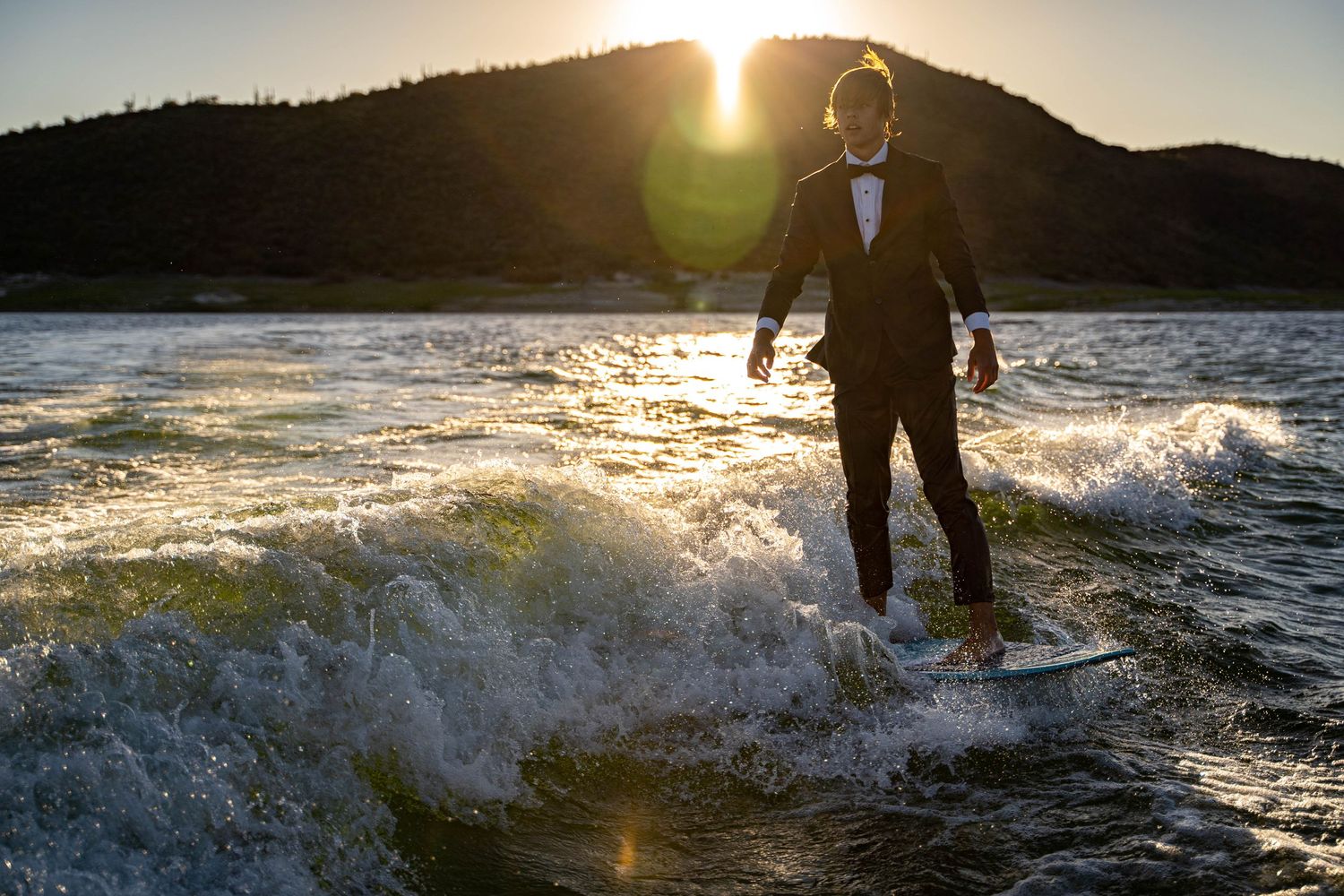 Silhouetted figure in formal attire surfing on waves at sunset with mountains in background.