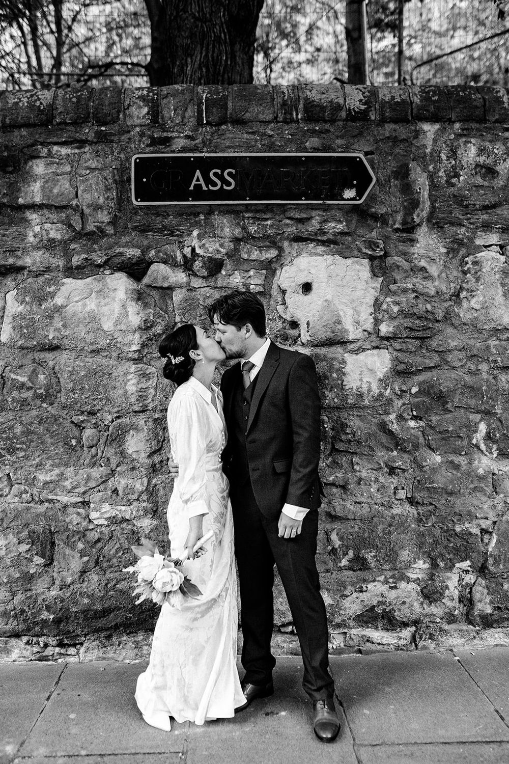 A black and white photo series of a couple in wedding attire posing against a stone wall.