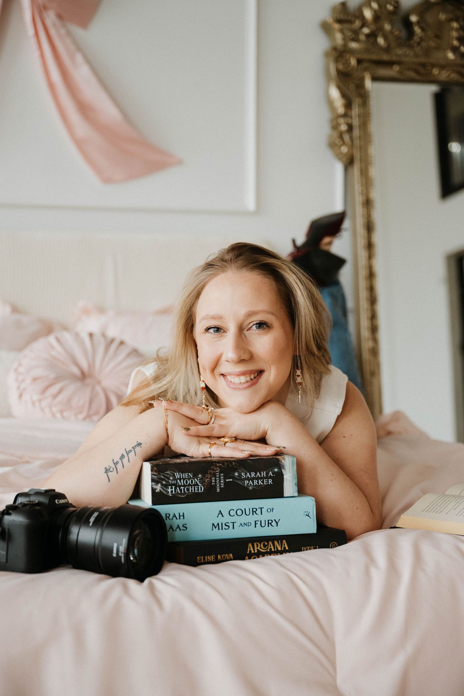 Smiling blonde woman lying on pink bed with stack of books and camera.