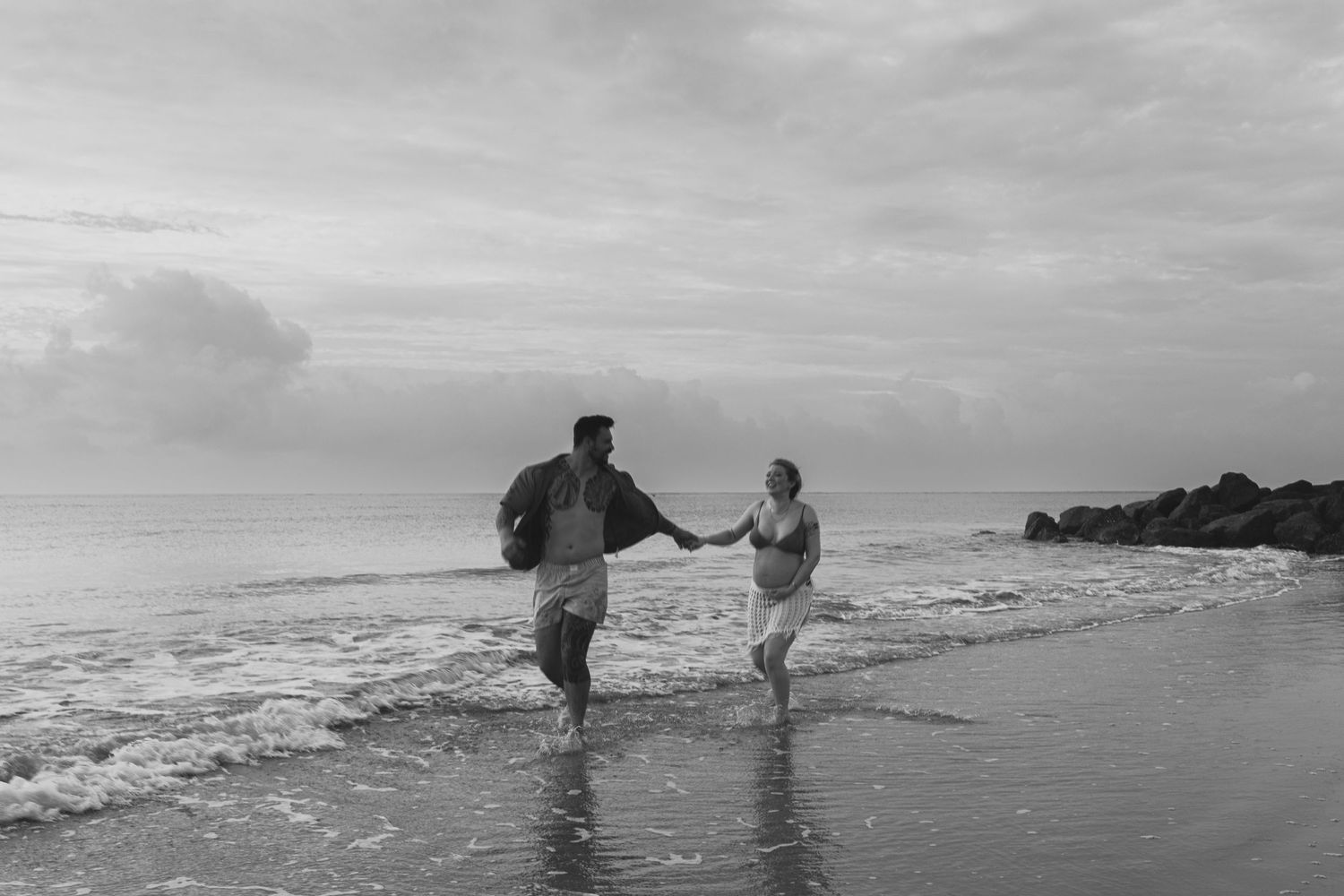 A couple runs joyfully along the beach shoreline in black and white photography.
