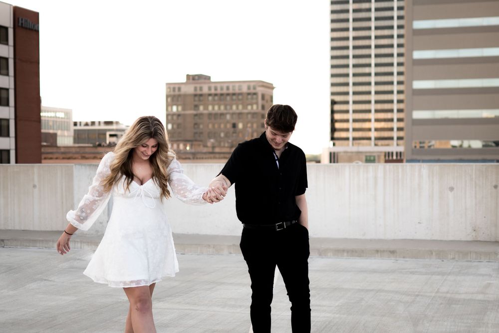 Couple in contrasting black and white outfits hold hands while walking on an urban rooftop with city buildings in background.
