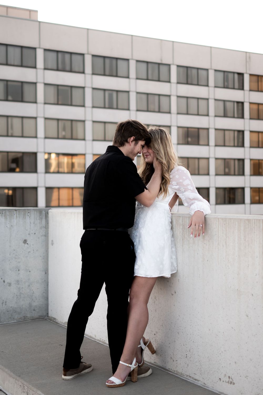 A romantic couple shares an intimate moment on a concrete balcony with an urban building backdrop.