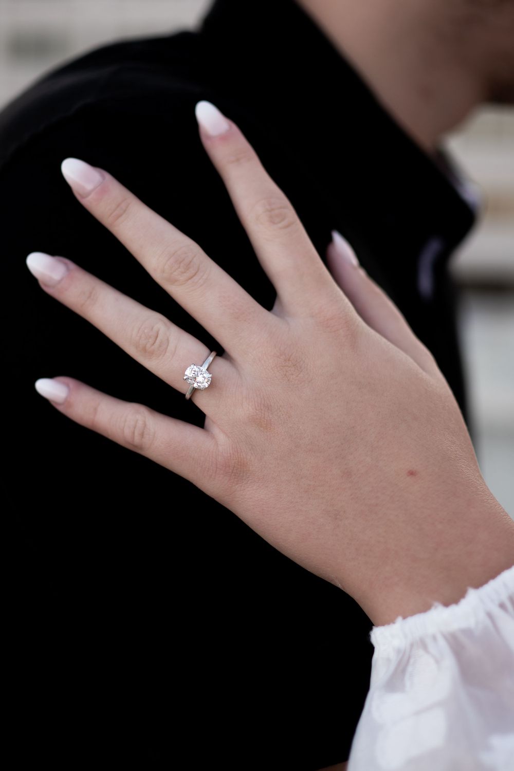 A hand wearing a white frilly sleeve displays a sparkling diamond engagement ring against a black background.