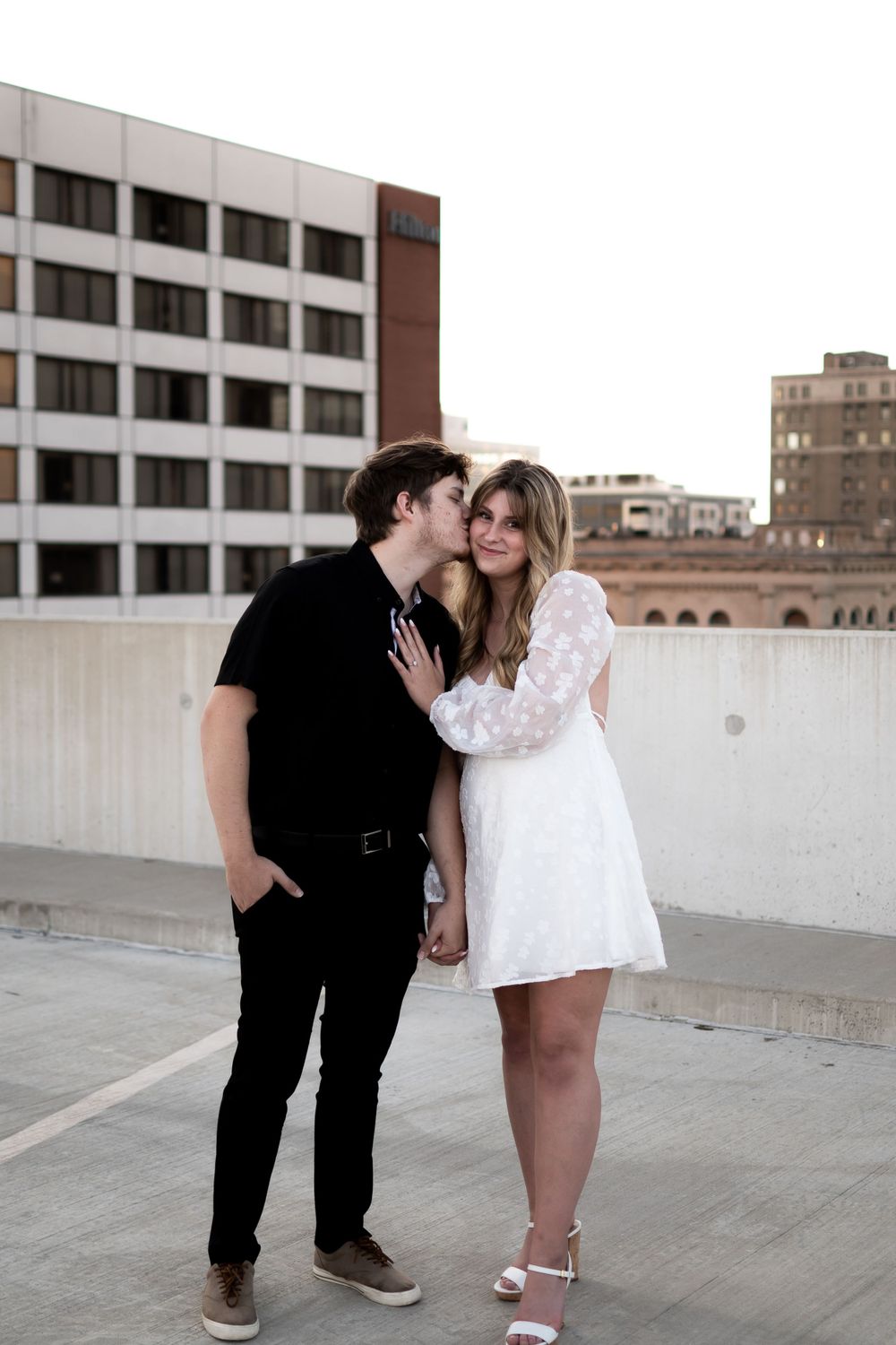 A romantic couple embrace on an urban rooftop with downtown buildings in the background at sunset.