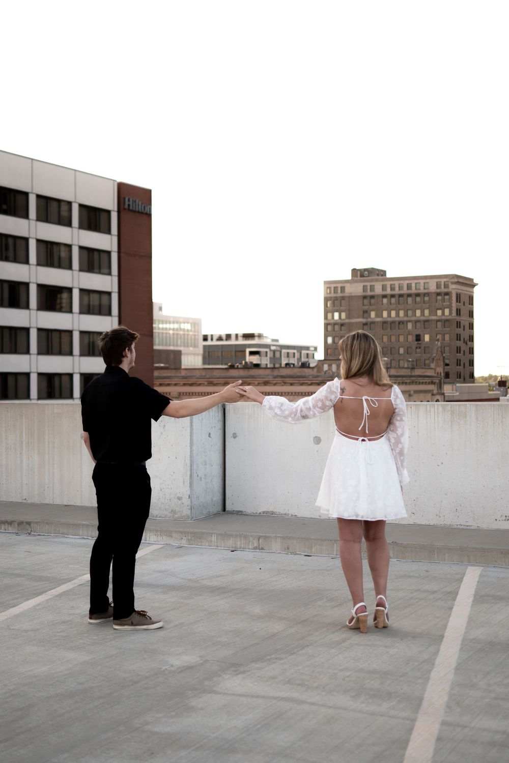 Two people hold hands while standing on an urban rooftop parking area overlooking city buildings at dusk.
