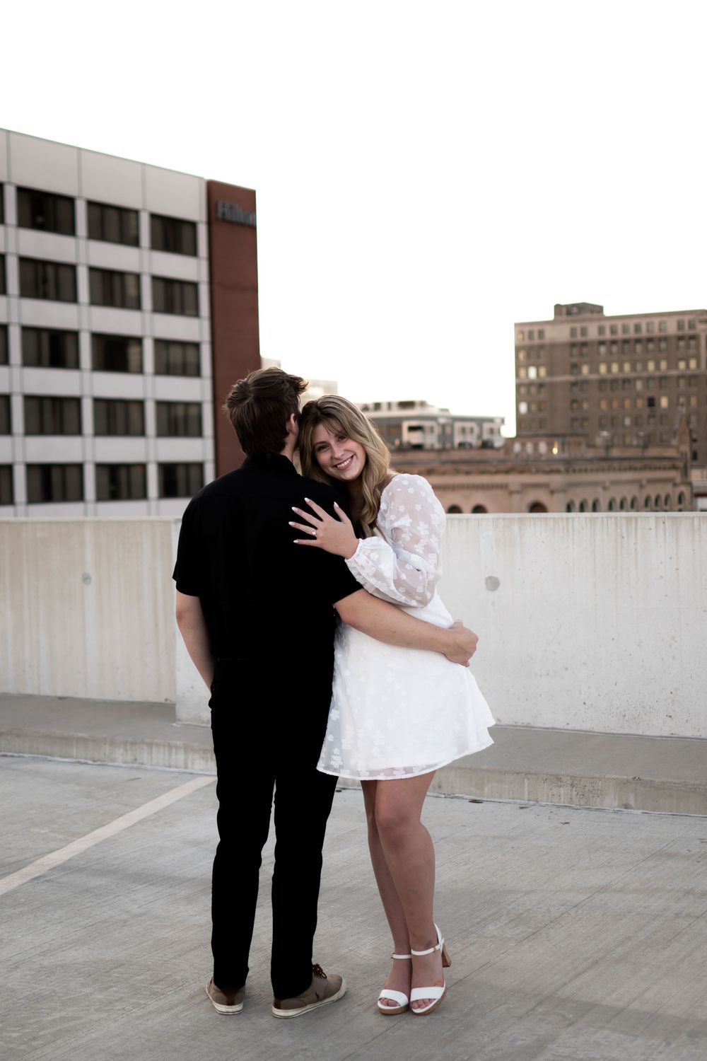 A couple embracing on an urban rooftop, with the person in white wearing a short dress and heels.