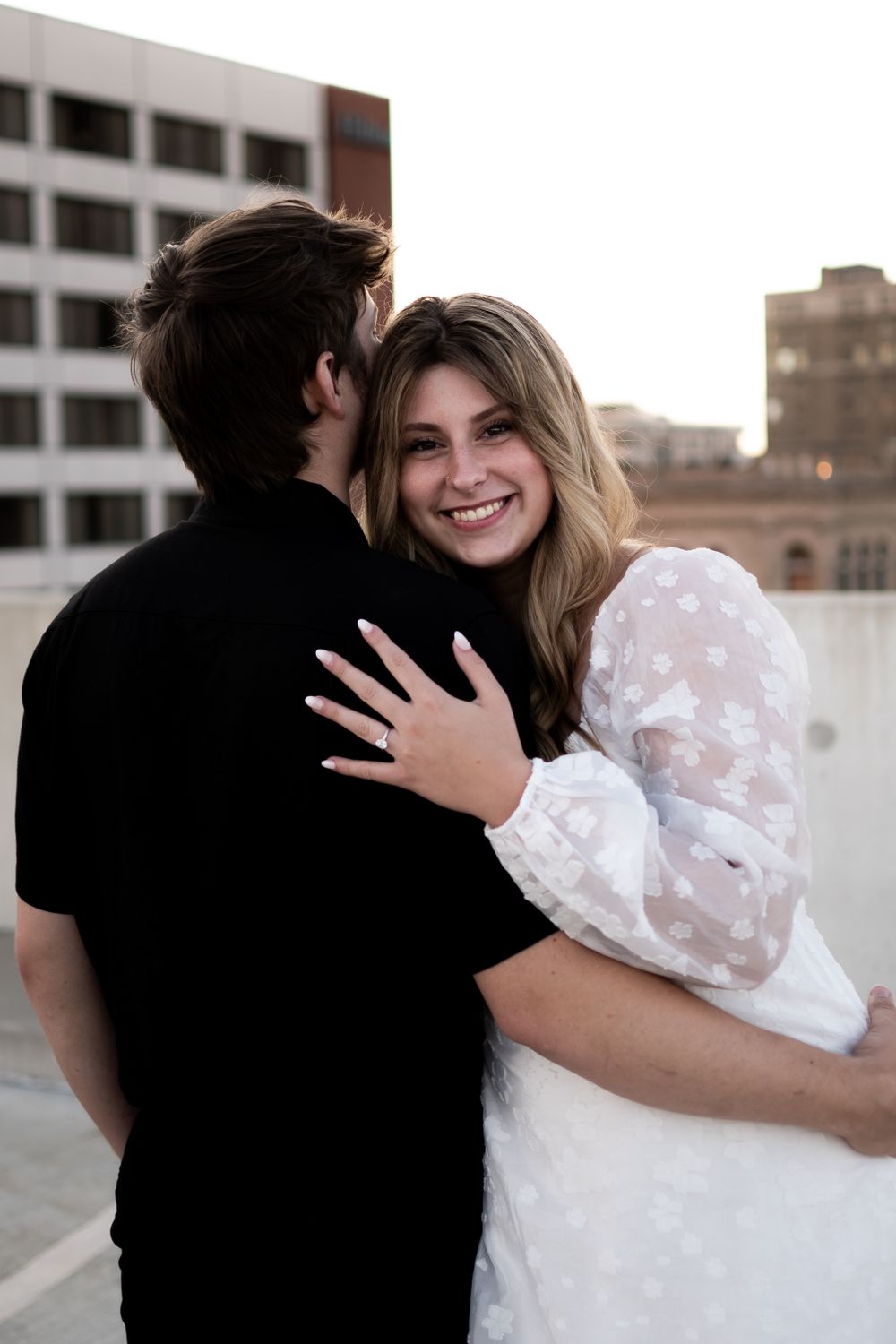 A couple embraces in a romantic moment on a rooftop with city buildings in the background as she shows her engagement ring.