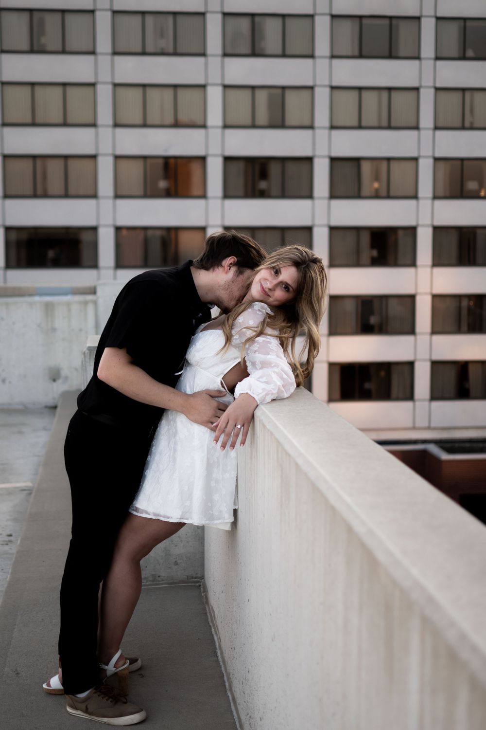 Romantic couple embraces on urban rooftop balcony with office building backdrop at dusk.