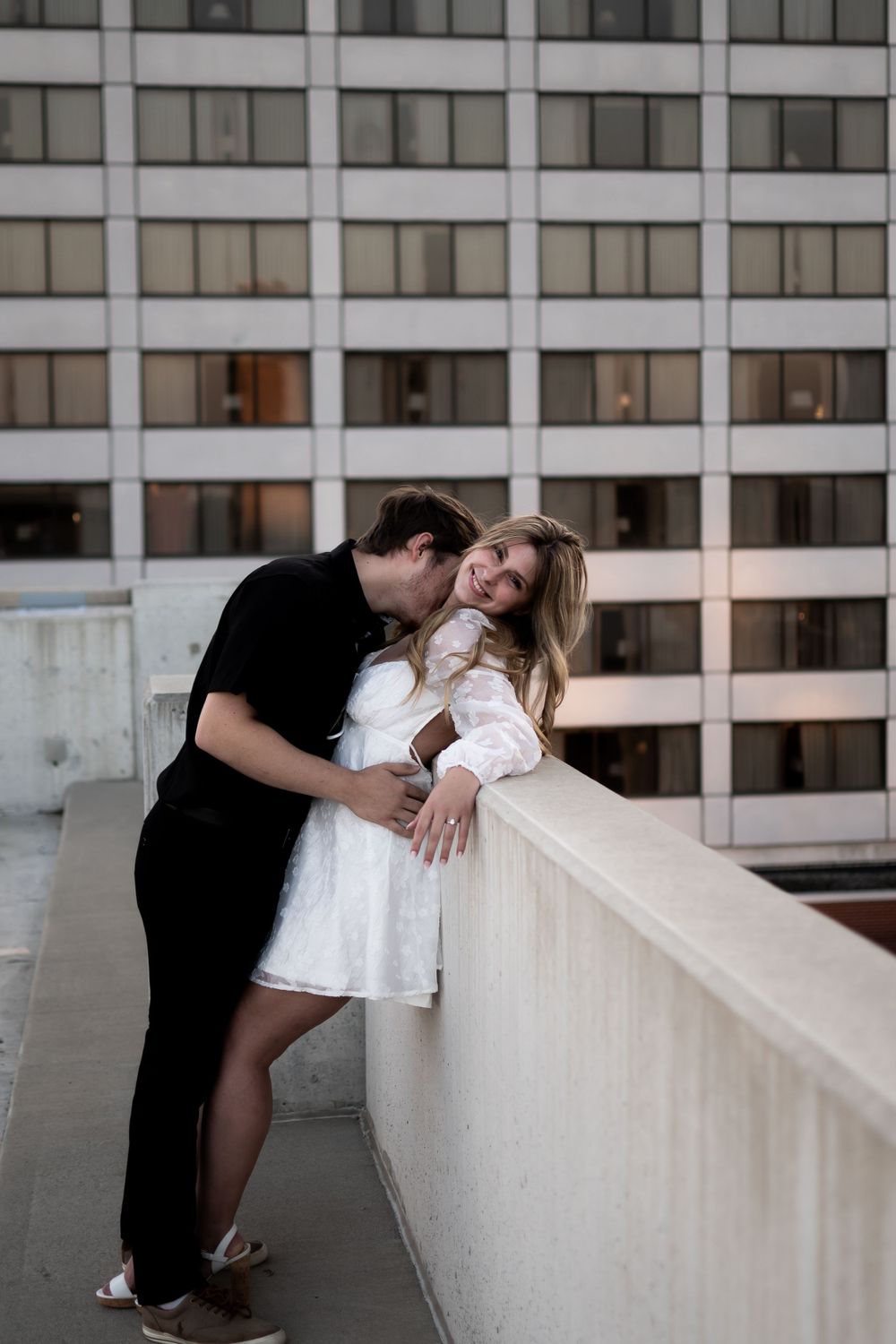A romantic couple embrace on an urban rooftop balcony with a modern office building facade in the background. Ashley Enfield Photography