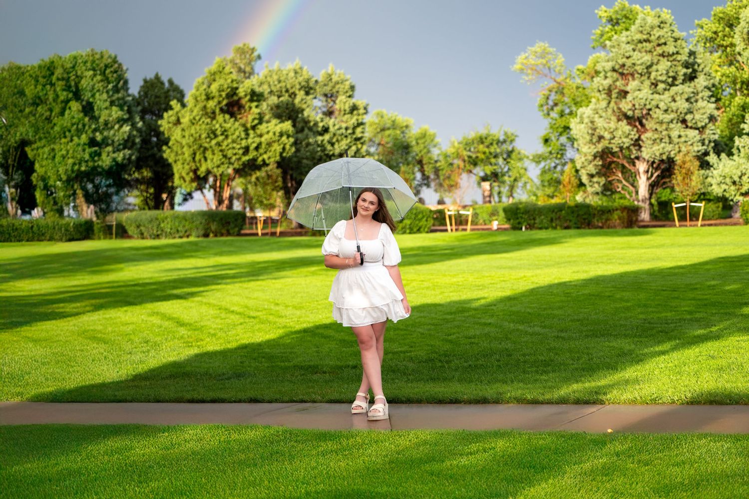 High school senior in white dress holds umbrella on green lawn with rainbow in background.