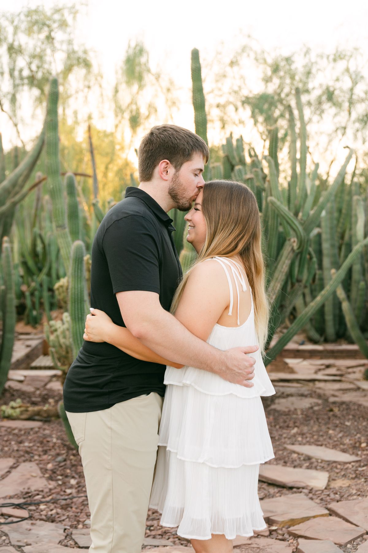 A man in a black shirt is kissing the forehead of a woman in a white dress. She's smiling and there's brown rocks and cactus behind them