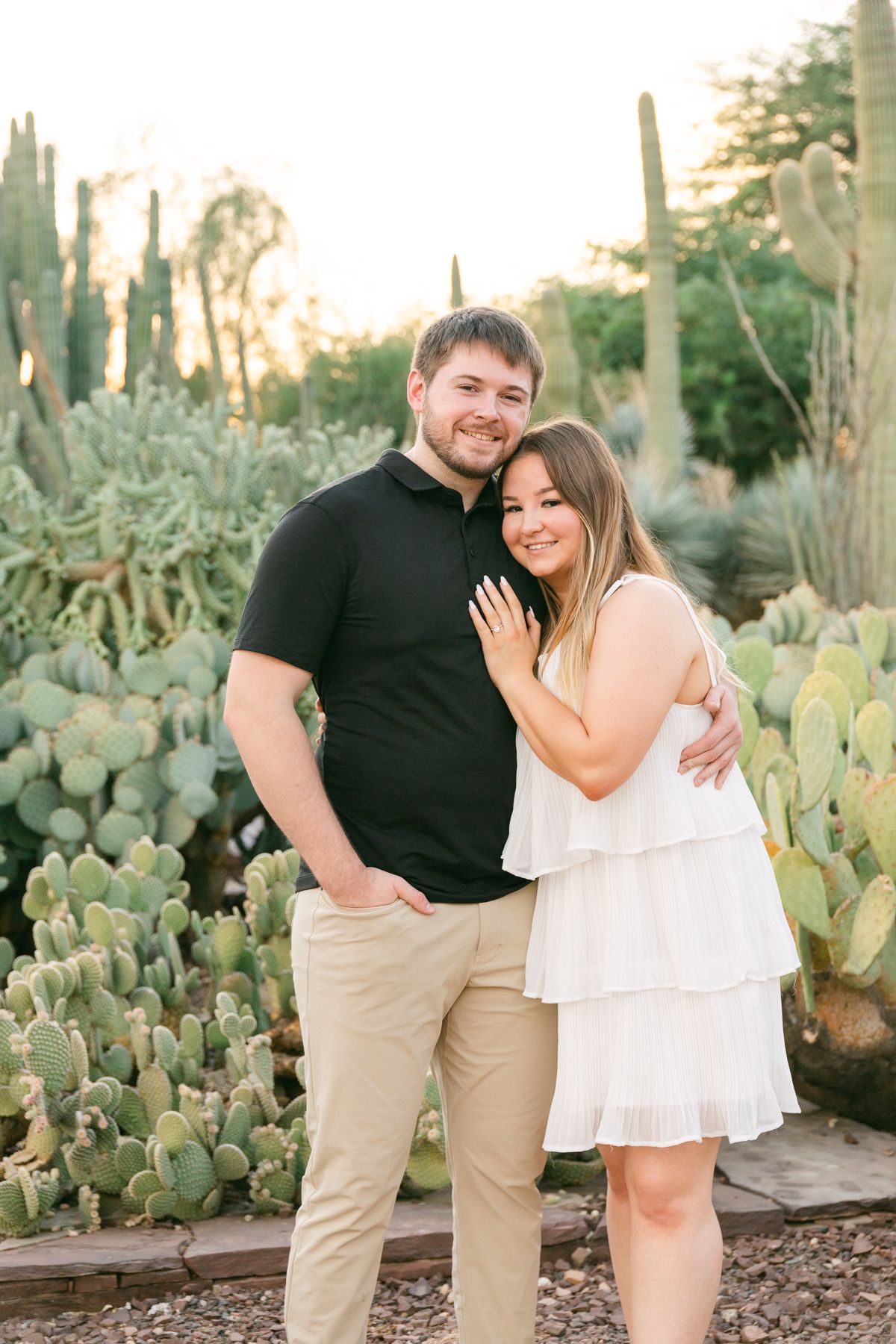 A man in a black shirt is holding a woman in a white dress. They're both smiling and there are cactus behind them.