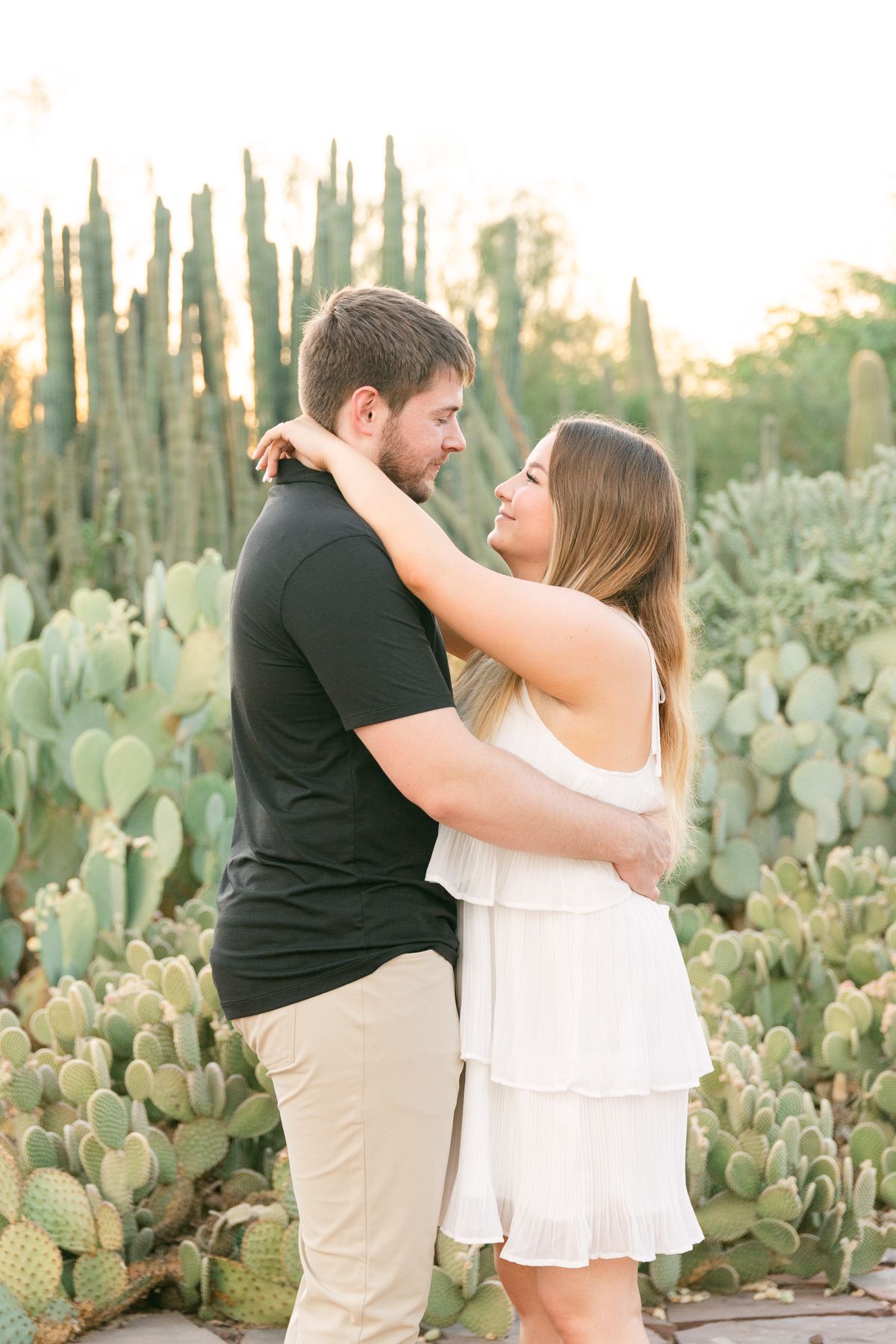 A man in a black shirt and a woman in a white dress have their arms around each other. They're smiling and there are cactus behind them.
