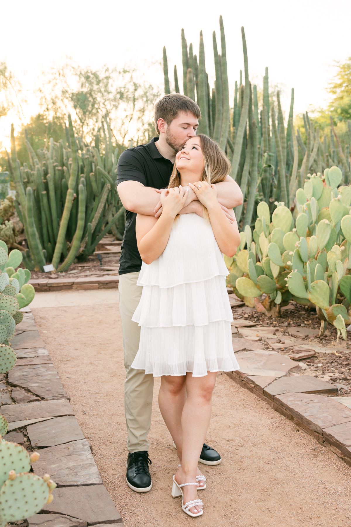 A man and a woman are embracing. She's in a white dress and he's in a black shirt kissing her head. There's a dirt path and cactus nearby