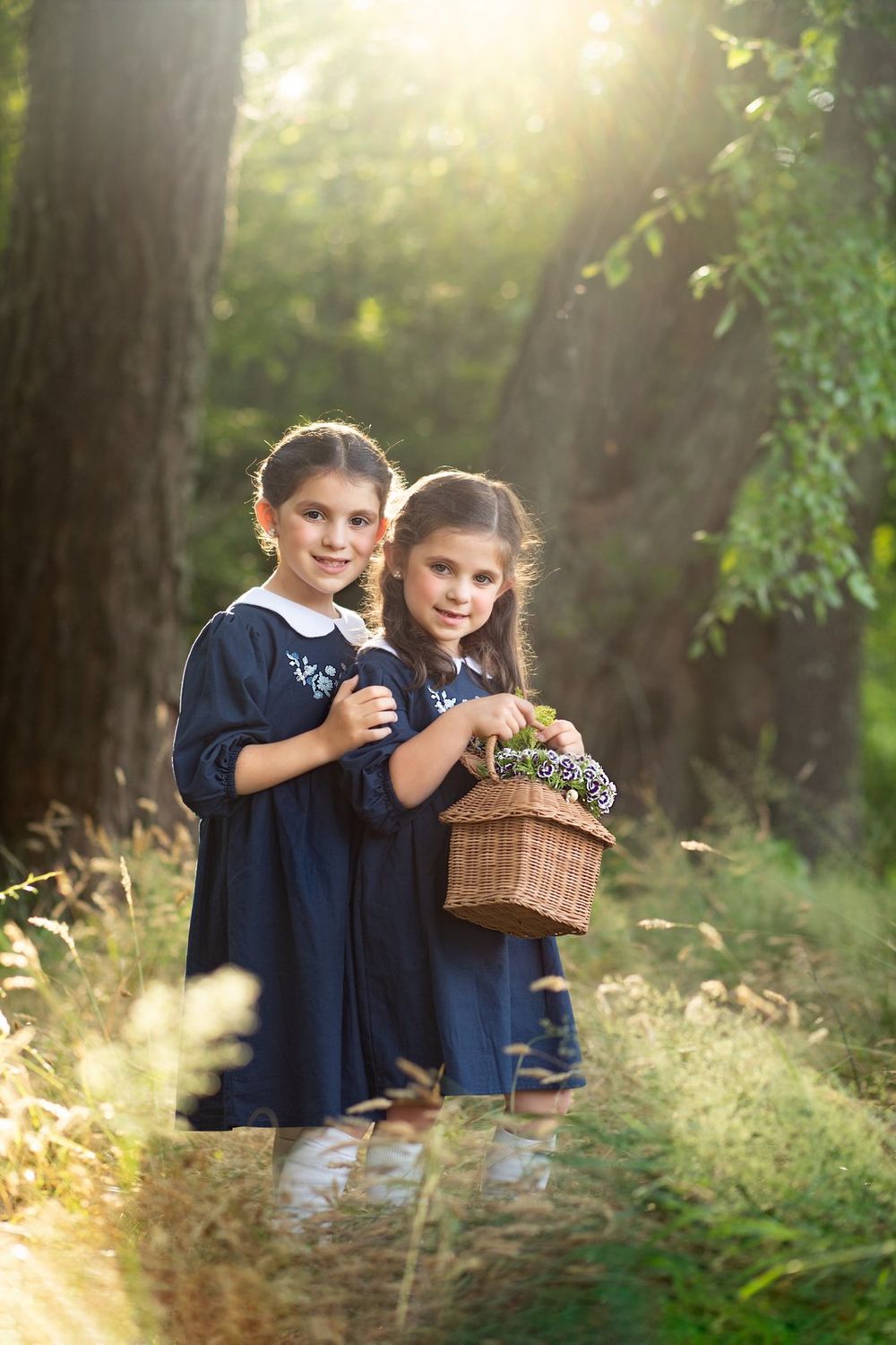 Two children in blue dresses holding a wicker basket stand in a sunlit forest path.