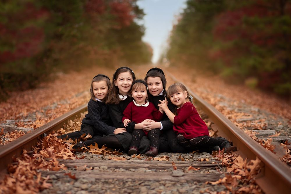 Five children in coordinated black and red outfits sit together on rustic train tracks covered in fall leaves.