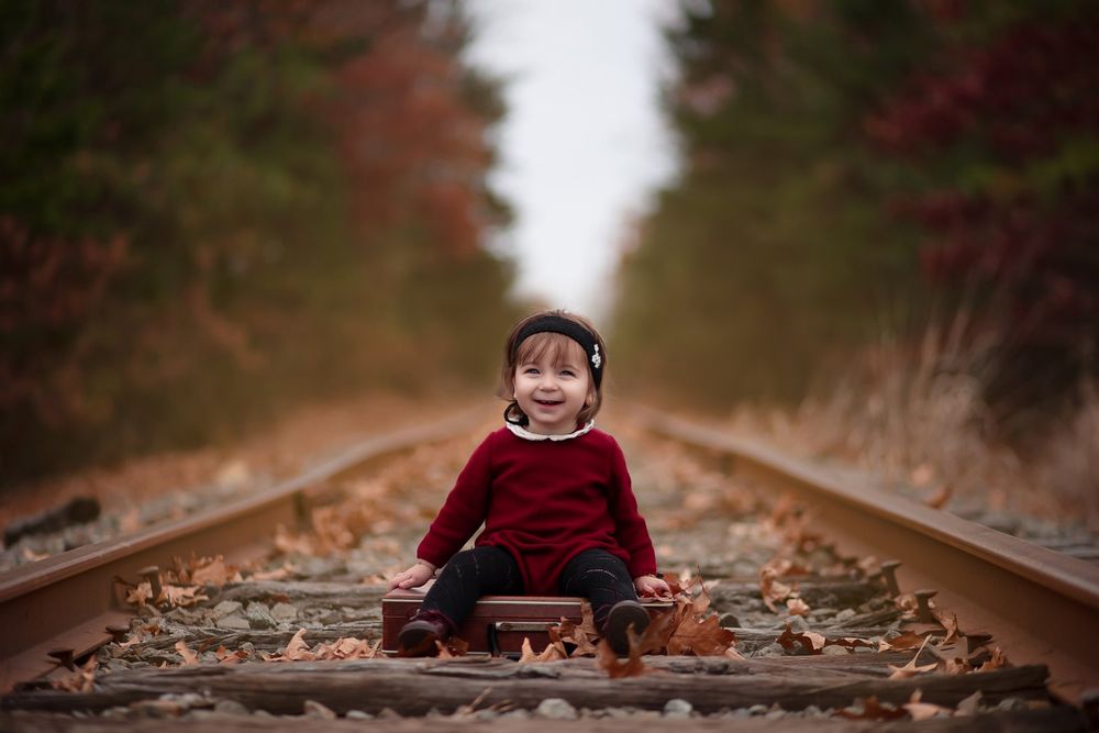 Young figure in red sweater sits playfully on train tracks surrounded by autumn foliage in a tunnel of trees.