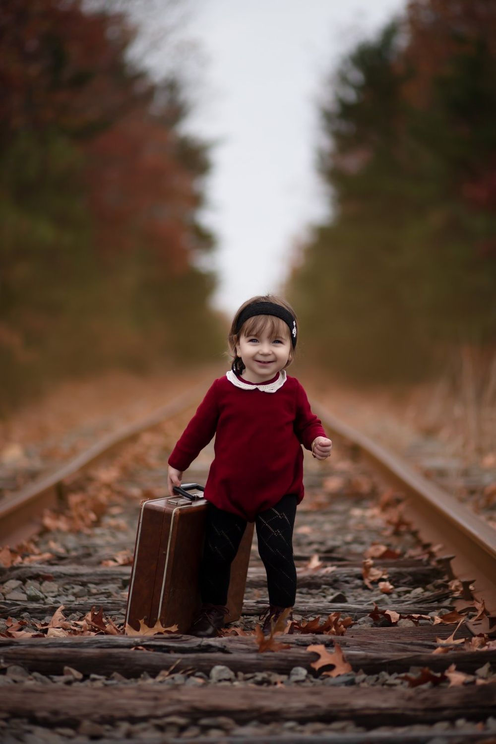 Small figure in red sweater carries vintage brown suitcase along autumn railroad tracks.