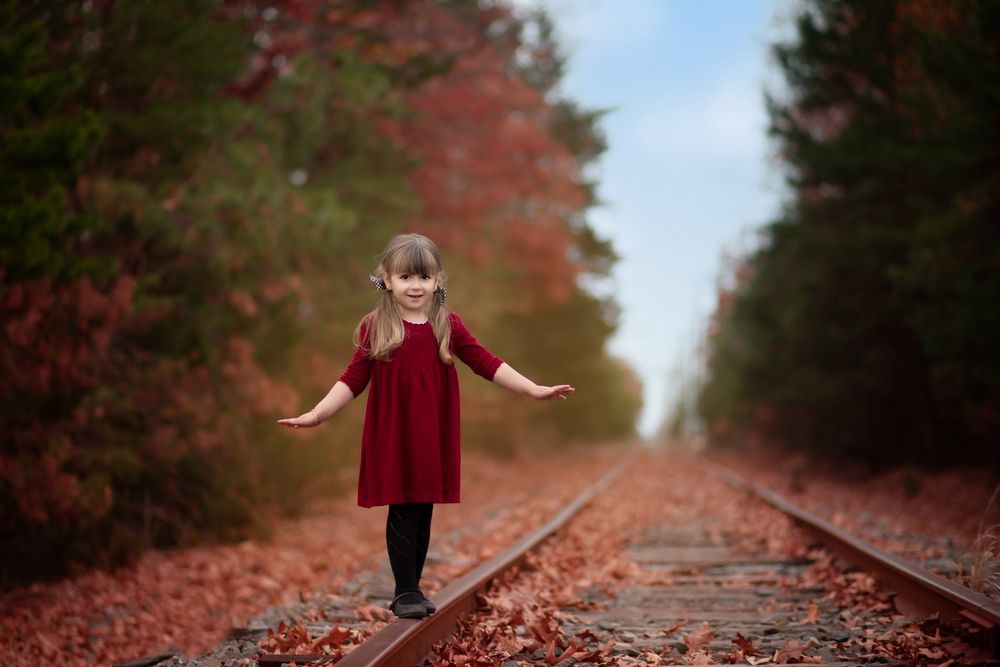 A child in a red velvet dress balances playfully on train tracks surrounded by autumn foliage.
