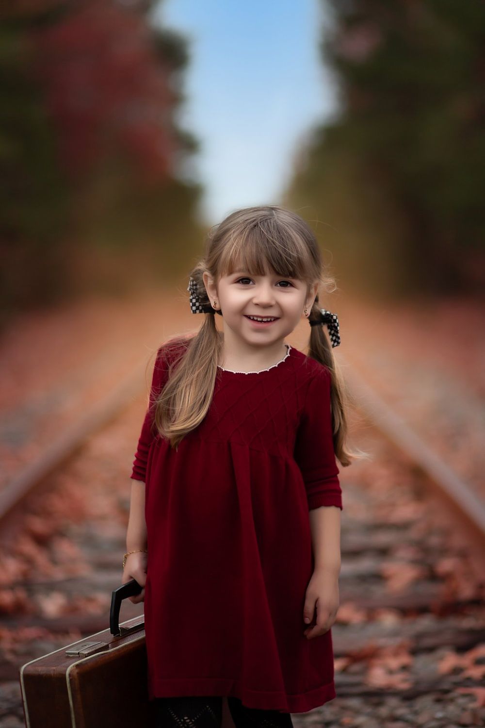 Little girl in red velvet dress stands on autumn train tracks holding vintage suitcase with leaves scattered around.