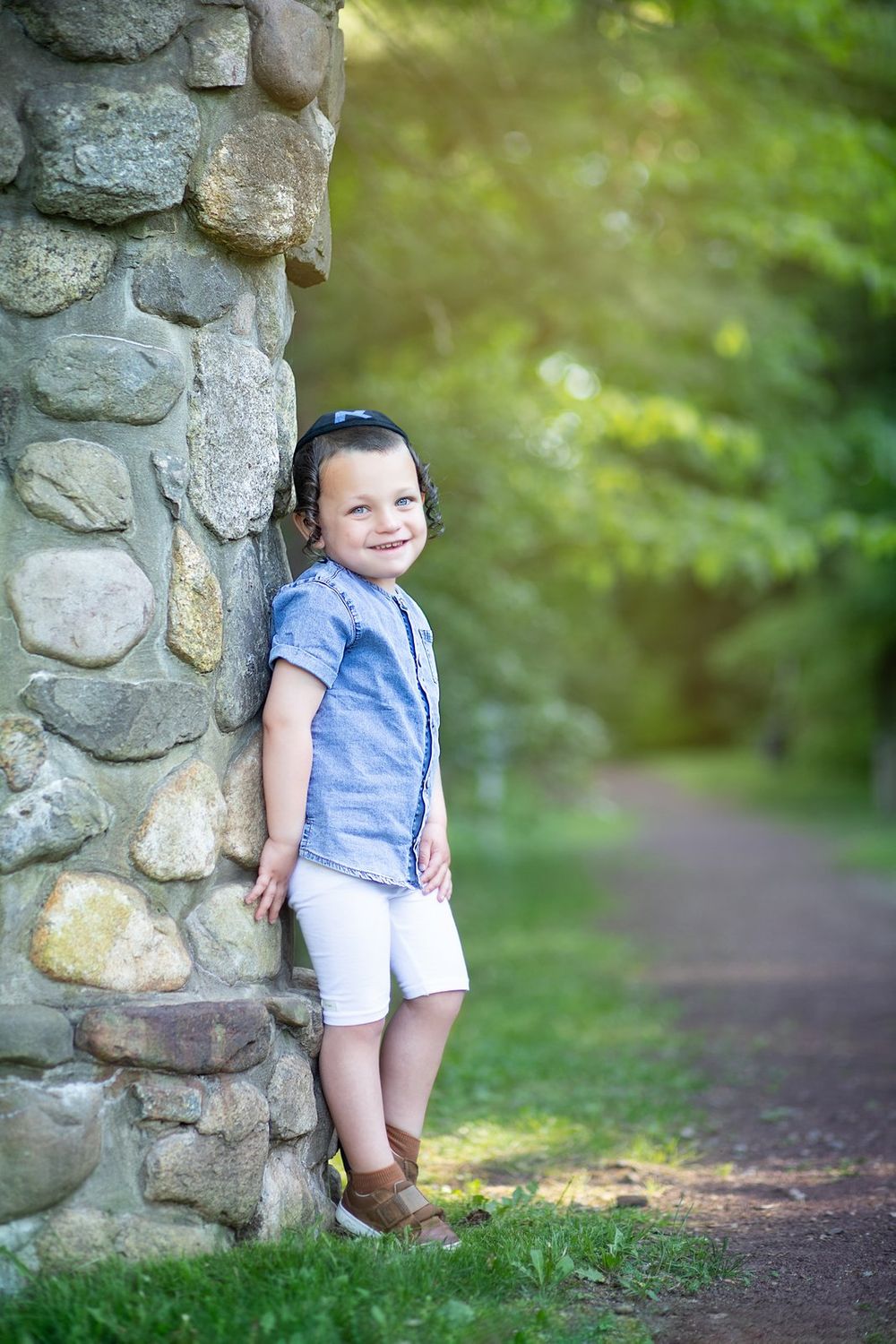 A young child in blue shirt and white shorts leans against a stone wall along a nature trail in dappled sunlight.