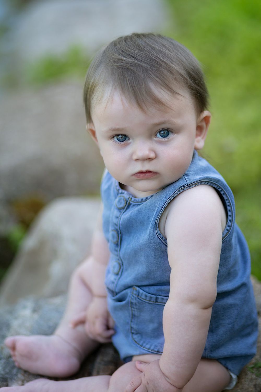 A young child in a denim romper sits outdoors on rocky ground against a blurred green background.