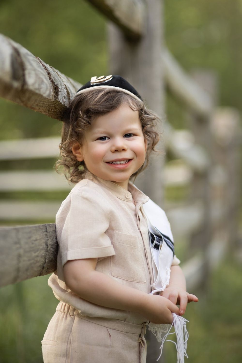A young child in beige clothing smiles while leaning against a wooden fence outdoors.