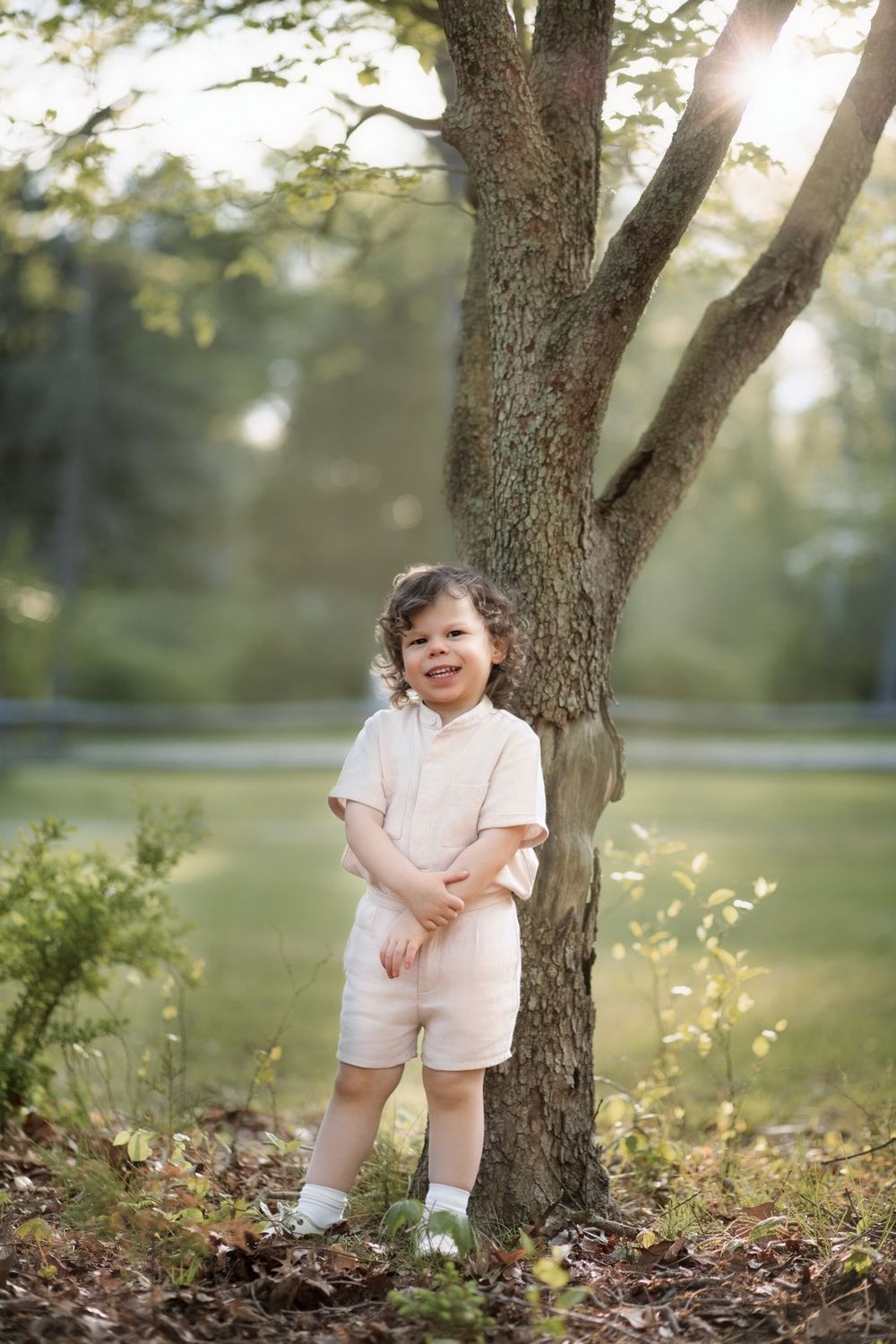 Child wearing white shorts outfit stands by tree trunk in dreamy sunlit garden setting.