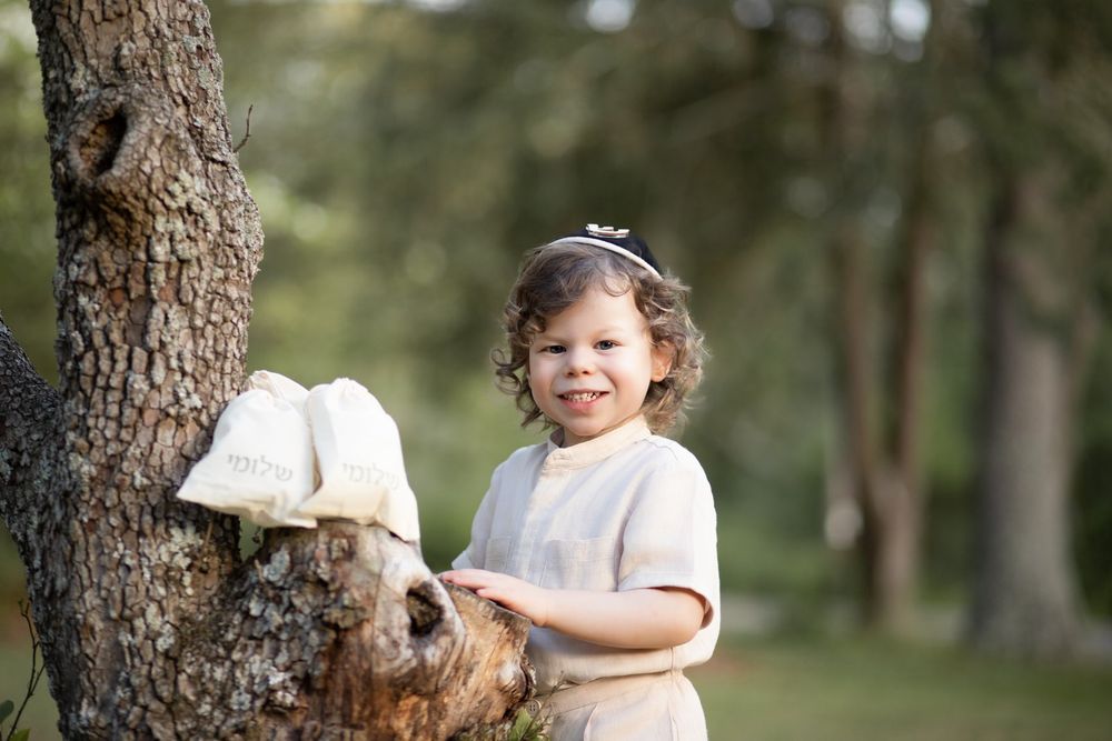 Young child in white stands by a tree with a stuffed animal toy in a woodland setting.