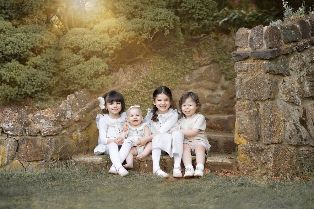 Children in matching white outfits sit together on rustic stone steps surrounded by lush greenery at sunset.