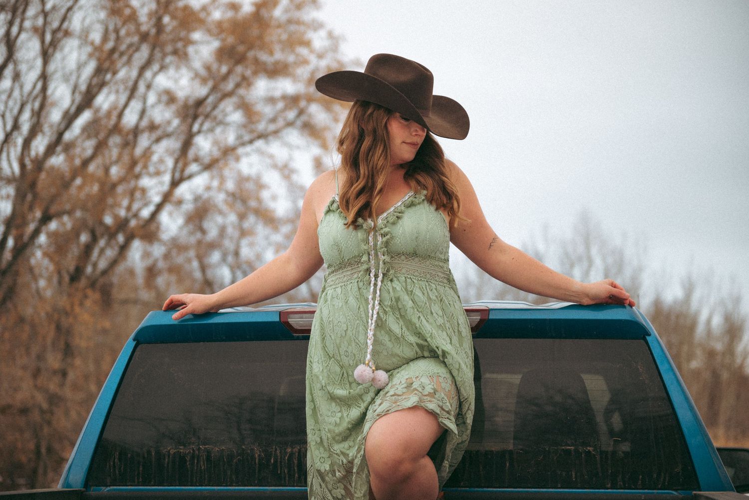 A woman in a green sundress and cowboy hat sits on the roof of a blue pickup truck at sunset.