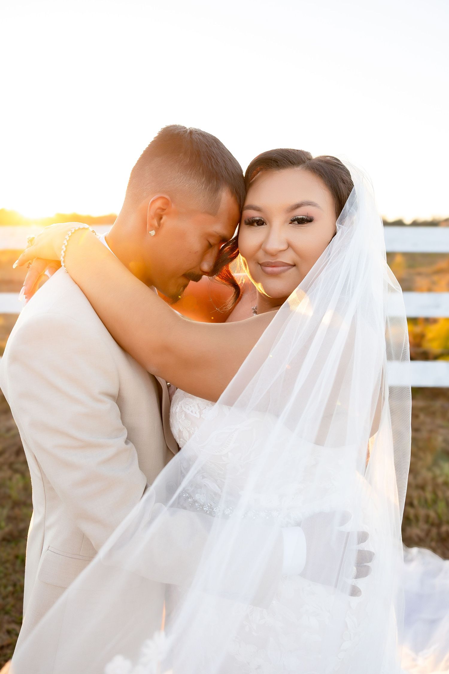 Interracial couple in wedding attire pose on a scenic gravel path lined with white fence and oak trees.