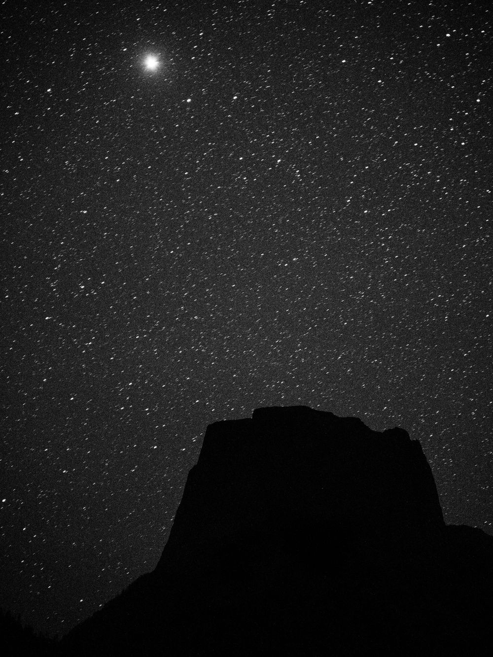 Black and white night sky filled with stars above a dark rocky mesa silhouette, with one bright star glowing prominently.