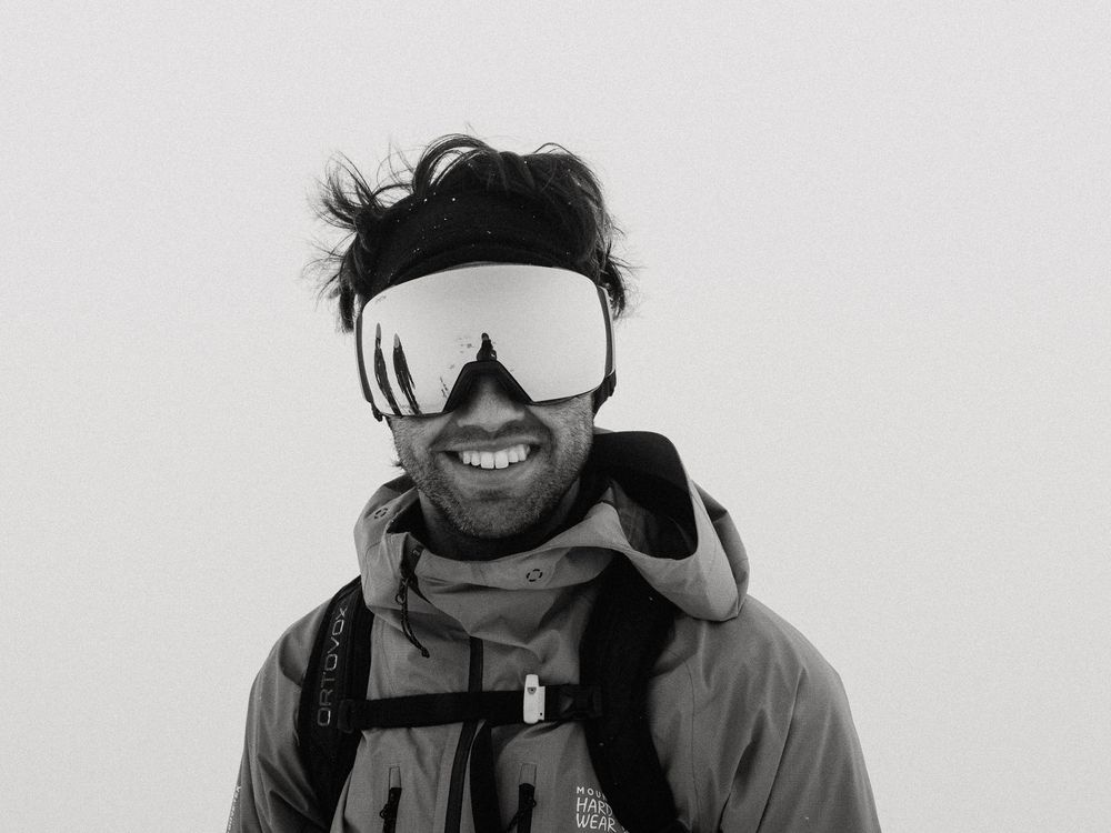 Smiling bearded man wearing ski goggles and jacket with backpack in black and white winter photo.