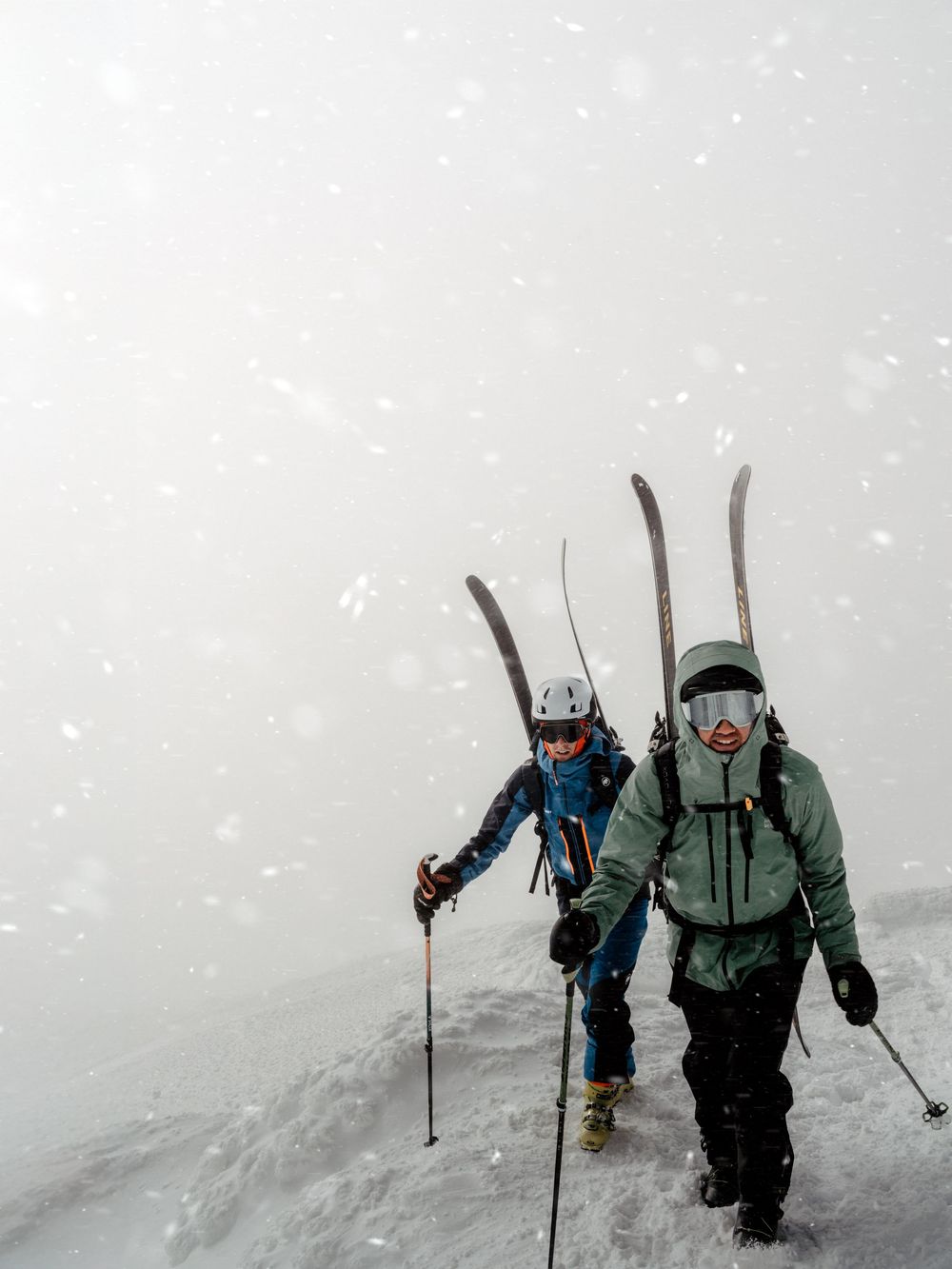 Two skiers carry skis on their backs while hiking uphill through a heavy snowstorm in winter mountain conditions.