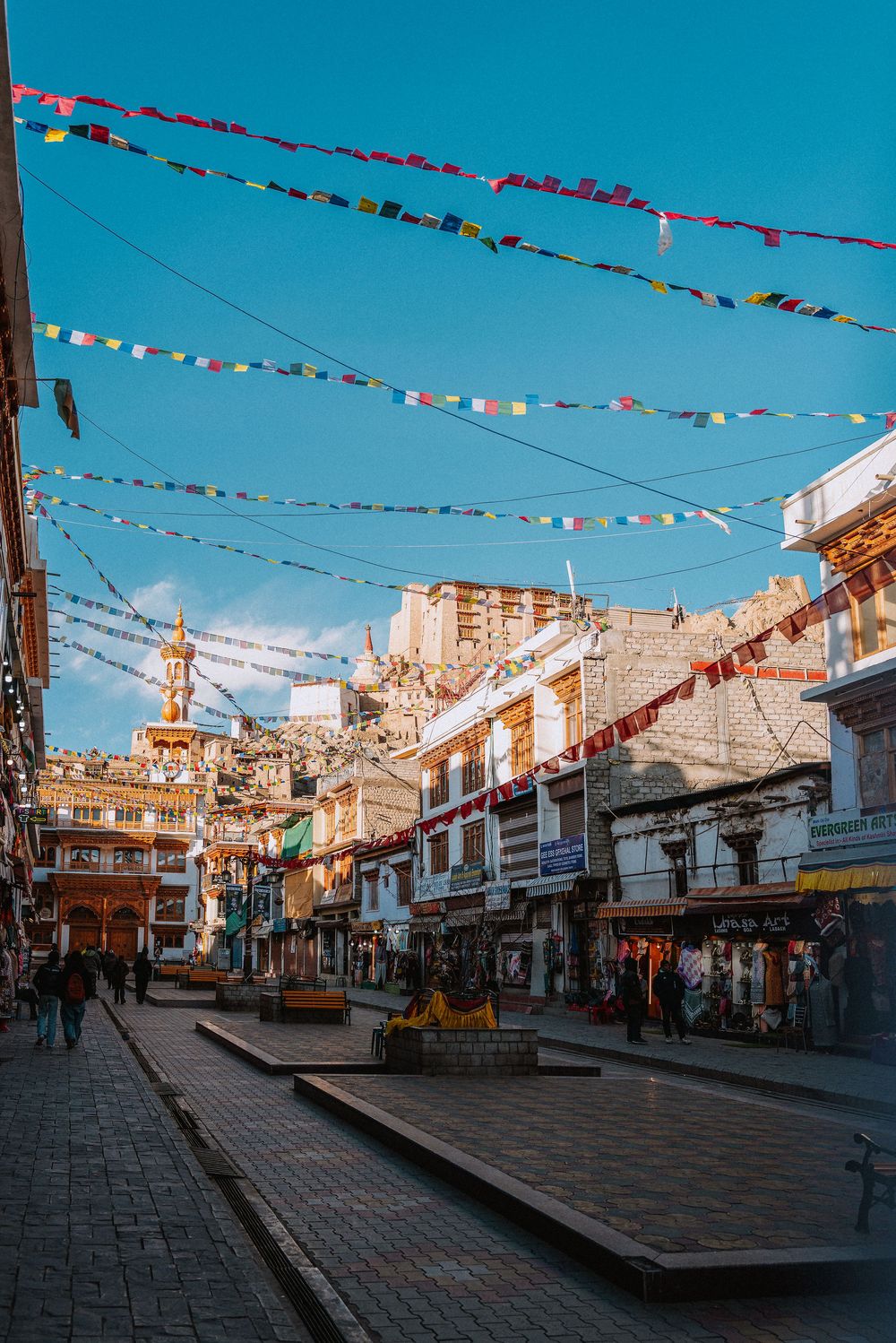 Colorful Buddhist prayer flags strung above a bustling market street in Leh, Ladakh, with historic buildings and clear blue sky.