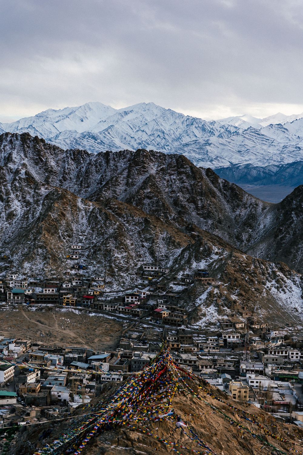 Aerial view of a Himalayan mountain town with colorful prayer flags, snow-capped peaks, and overcast skies in Ladakh, India.