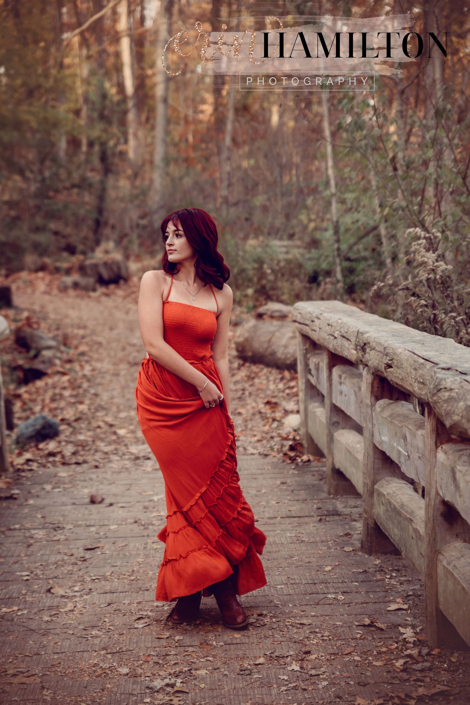 A flowing red dress catches the autumn breeze as someone poses on a rustic wooden bridge in a forest setting.