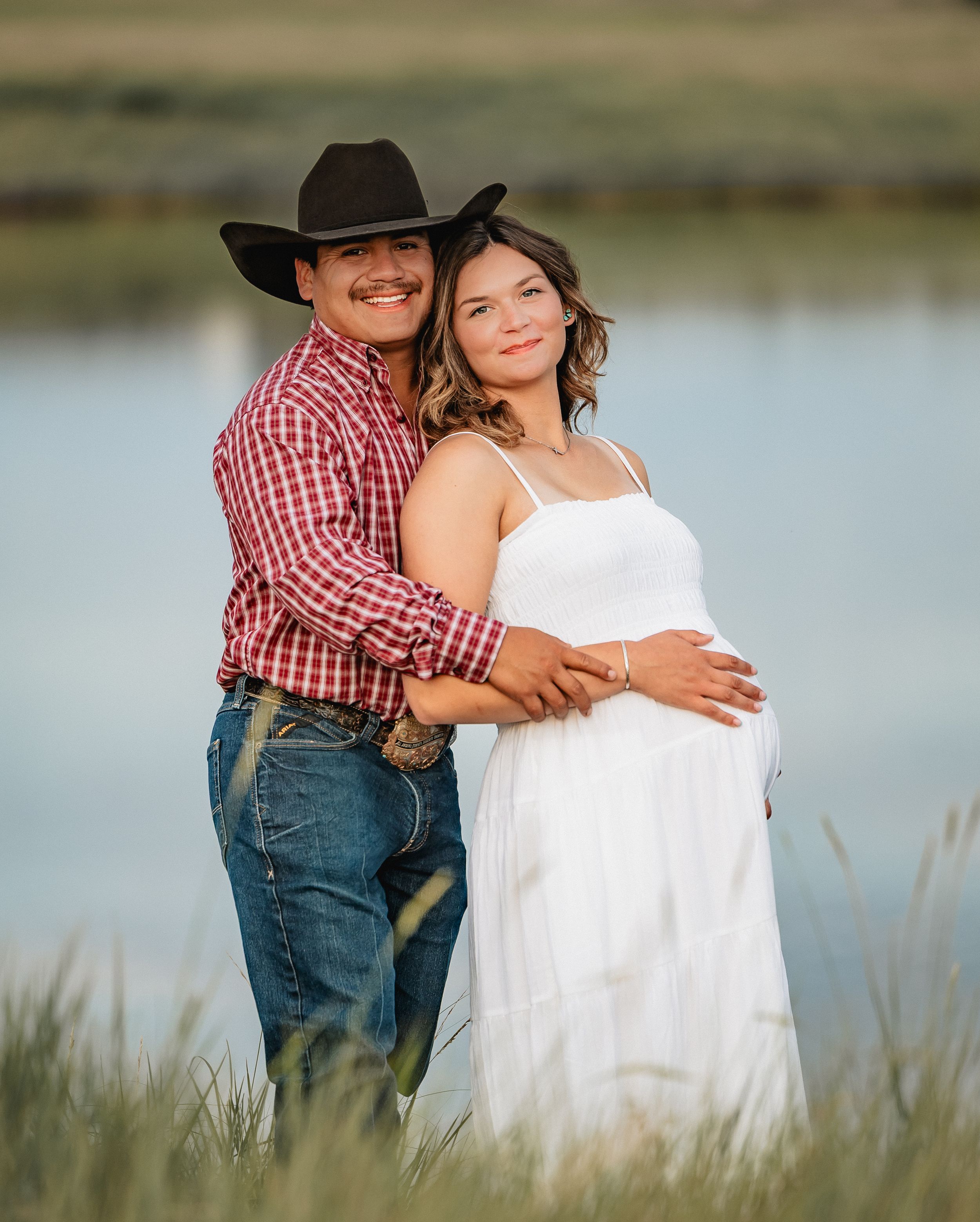 Couple poses outdoors near a lake, man in cowboy hat and plaid shirt embraces pregnant woman in white dress.