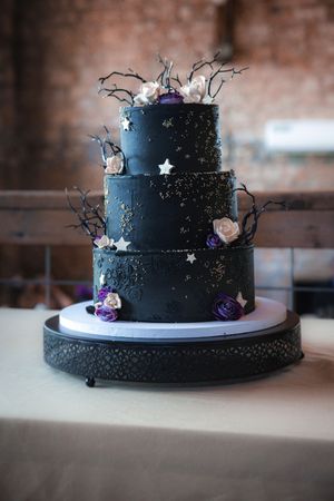A three-tiered black wedding cake decorated with silver stars, purple flowers and twisted branches on a decorative stand.