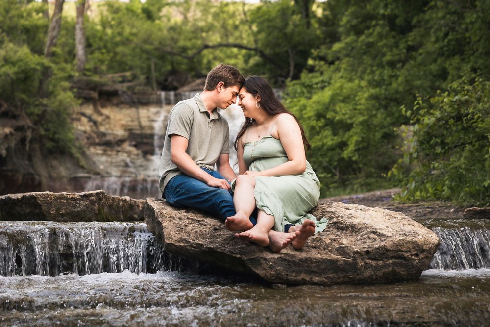 Couple sitting close on rocks by a waterfall surrounded by lush greenery in a romantic outdoor setting.
