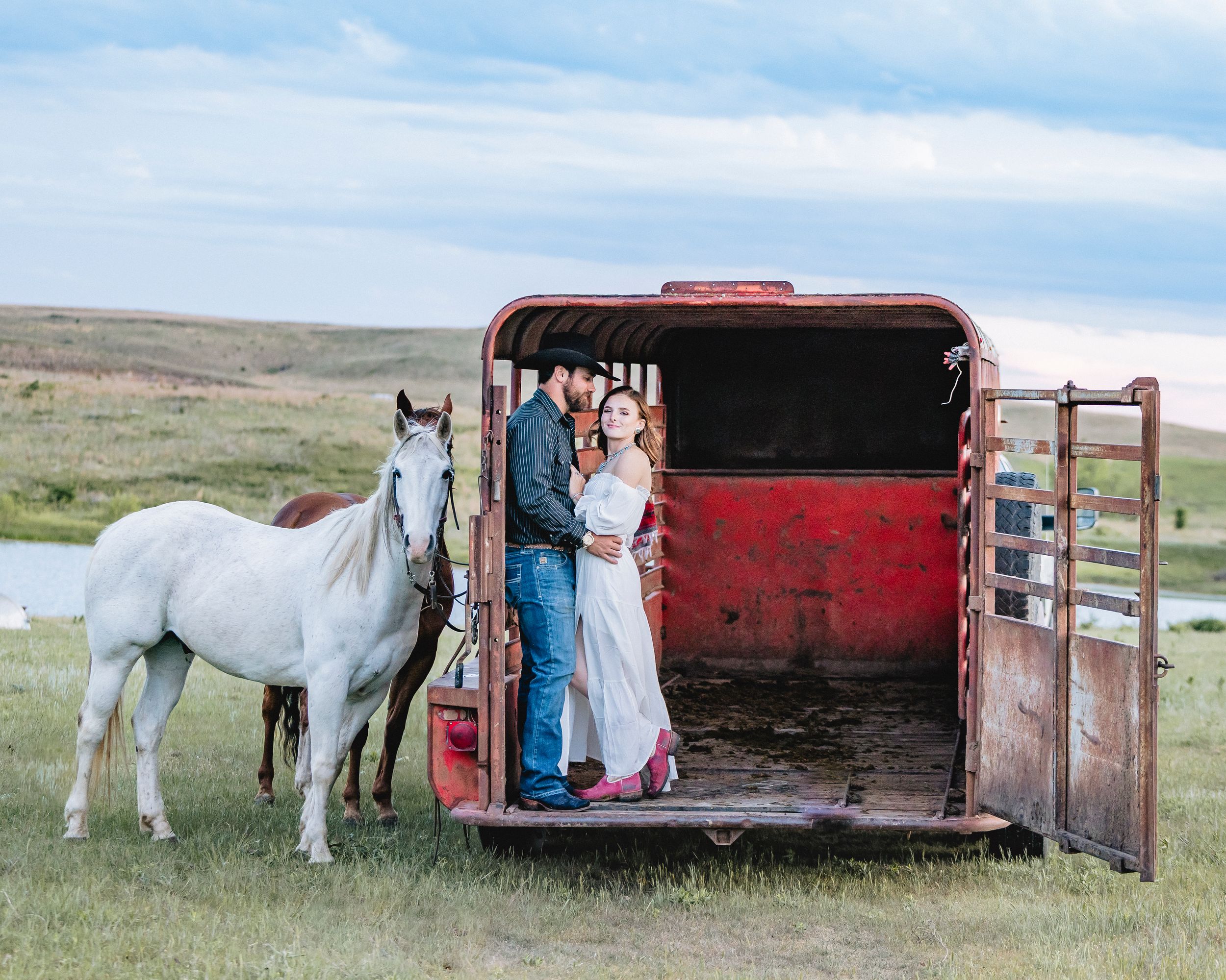 Couple embracing in red horse trailer with white horse nearby in green pasture under cloudy sky.